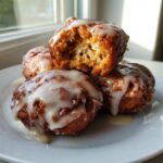 Close-up of three glazed apple fritter mounds stacked on a white plate, one broken open showing the soft interior.