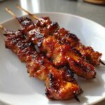 Three glossy, caramelized chicken skewers resting on a white plate, showing char marks from grilling.