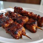 Four glistening, caramelized chicken skewers with a dark, sticky glaze resting on a white plate.