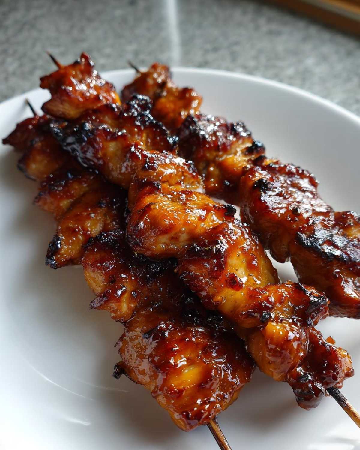 Close-up of three glossy, glazed chicken skewers resting on a white plate, featuring caramelized edges.