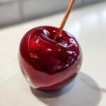Close-up of a perfectly glossy, deep red candy apple on a white surface with a wooden stick inserted.
