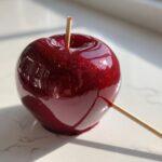 A single, perfectly glossy, deep red candy apple with visible glitter resting on a white marble surface.