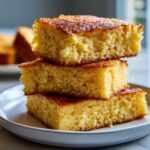 Three square pieces of golden fried cornbread stacked on a white plate, showing a moist, crumbly texture.