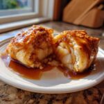 A warm apple dumpling cut in half, showing soft apple slices inside, drizzled with rich caramel sauce.