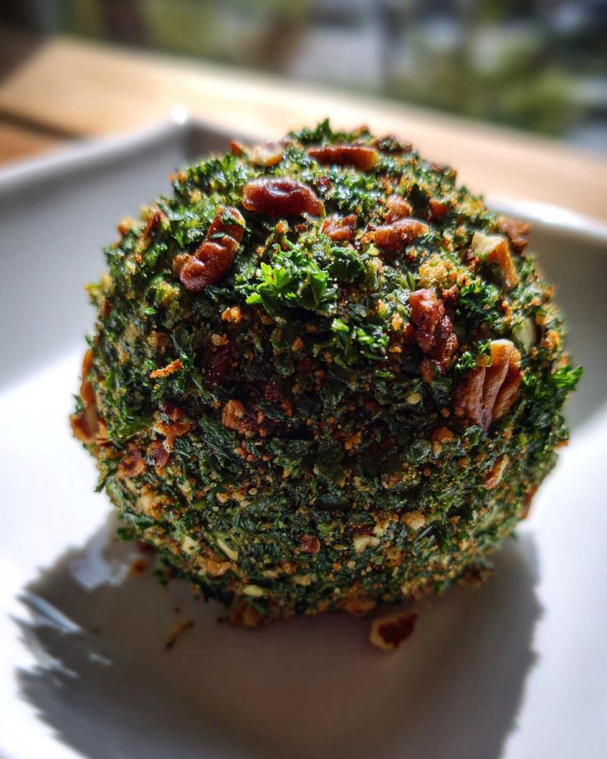 A close-up of a perfectly formed cheese ball recipe coated heavily in green herbs and chopped pecans.