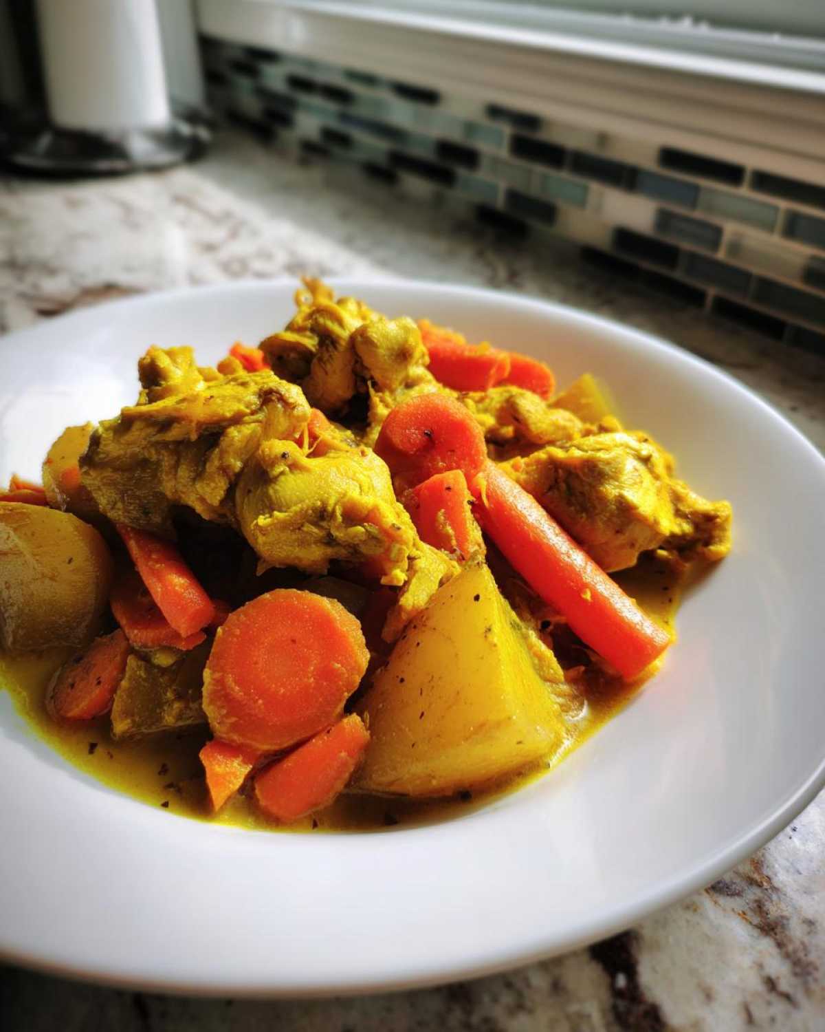 A close-up of a white plate holding a serving of vibrant yellow Jamaican curry chicken mixed with chunks of potato and bright orange carrots.