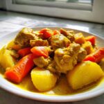 Close-up of a white plate filled with rich, yellow Jamaican curry chicken, potatoes, and bright orange carrots.