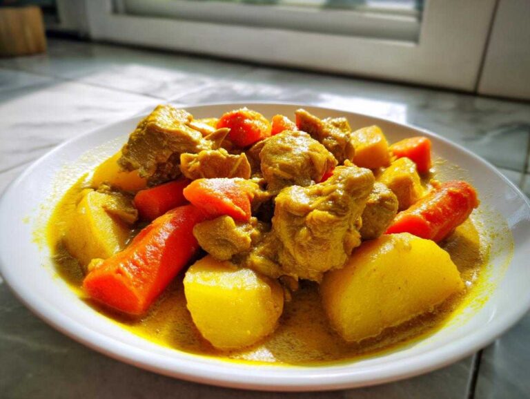 Close-up of a white plate filled with rich, yellow Jamaican curry chicken, potatoes, and bright orange carrots.