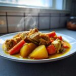 Close-up of steaming Jamaican curry chicken pieces served with large chunks of potato and bright orange carrots in a rich yellow sauce.