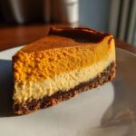 A perfect slice of layered pumpkin cheesecake featuring a dark crust, a light cream cheese layer, and a vibrant orange pumpkin top layer.