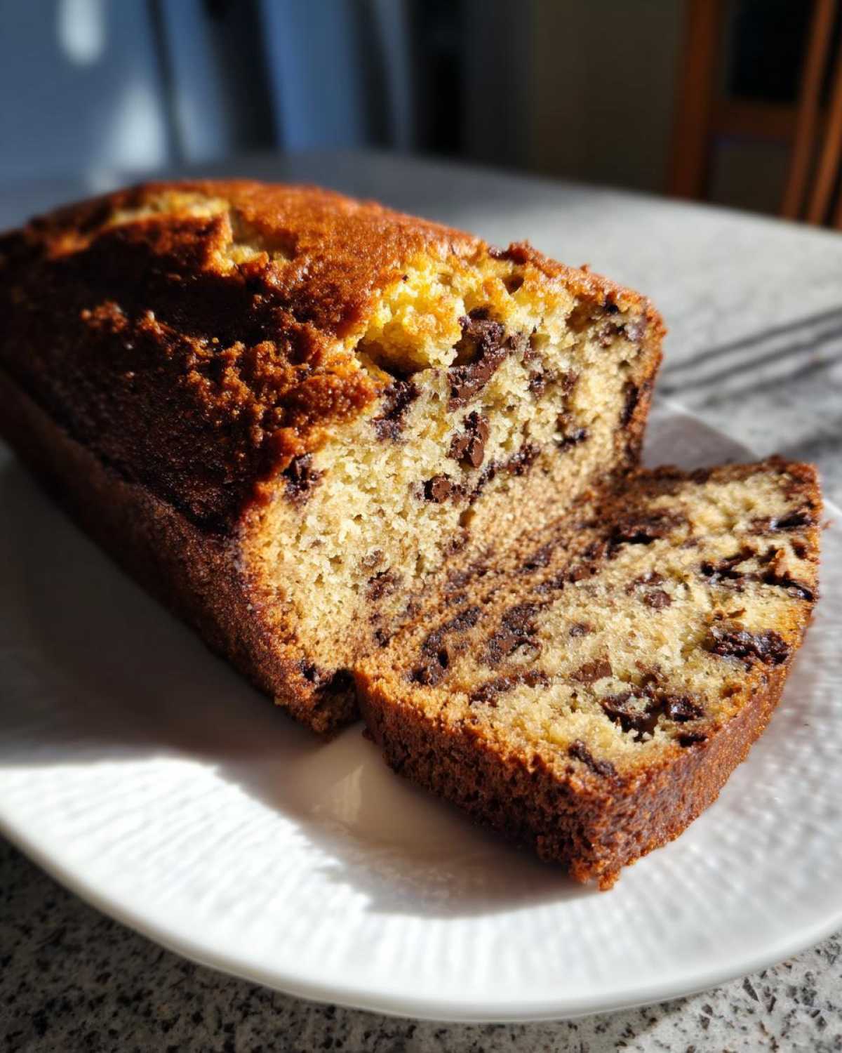 A freshly baked chocolate chip banana bread recipe loaf, partially sliced, sitting on a white plate.