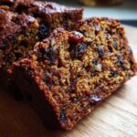 Close-up of two moist slices of dark fruit cake recipe packed with raisins and nuts on a wooden board.
