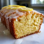 A close-up of a moist lemon loaf cake, drizzled with white icing and topped with thin lemon slices.