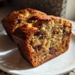 Close-up of a moist mini banana bread loaf cut in half, showing chocolate chips inside, on a white plate.