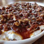 Close-up of a white dish filled with creamy pecan pie dip, topped with glossy caramel sauce and chopped pecans.