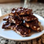 A stack of broken pieces of rich, dark chocolate peanut butter pretzel bark piled on a white plate.