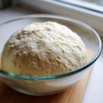 A large, risen ball of pizza dough recipe resting in a clear glass bowl, showing texture after proofing.