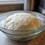 A large ball of risen pizza dough recipe resting in a clear glass bowl on a kitchen counter.