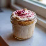 A single serving Protein Cheesecake layered in a glass jar with a graham cracker crust and raspberry swirl topping.