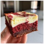 A close-up shot of a single, thick square of red velvet cheesecake brownies held by a hand.