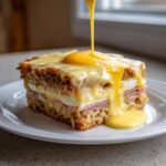 A square serving of eggs benedict casserole topped with ham, egg, and rich Hollandaise sauce being poured over it.