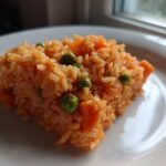 A square serving of moist, orange-hued Spanish rice mixed with bright green peas and diced carrots on a white plate.