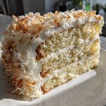 A moist slice of coconut cake recipe layered with white frosting and covered in toasted coconut flakes.