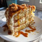 A tall slice of caramel crunch cake featuring multiple layers of cake, cream frosting, and dripping caramel sauce.