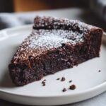 A close-up of a dense, dark slice of flourless chocolate torte dusted with powdered sugar on a light plate.