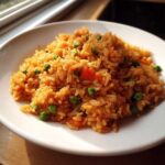 A close-up serving of fluffy, orange-colored Spanish rice mixed with bright green peas and diced carrots on a white plate.
