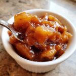 A spoonful of thick, glossy, spiced apple pie filling being lifted from a small white bowl.