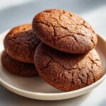 A close-up stack of three dark, rich soft gingerbread cookies with cracked tops on a light plate.