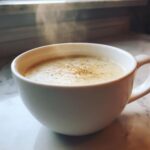 A close-up of steaming hot cauliflower soup, seasoned with pepper, served in a white mug on a marble surface.