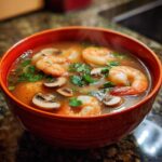 Close-up of a steaming orange bowl filled with flavorful tom yum soup featuring large shrimp and sliced mushrooms.