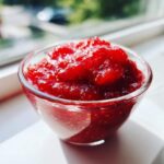 A close-up of bright red, chunky strawberry freezer jam served in a small clear glass bowl near a window.