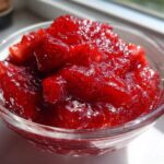 Close-up of vibrant, chunky strawberry freezer jam glistening in a small clear glass bowl near a window.