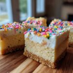 Four square slices of sugar cookie cheesecake bars topped with white frosting and colorful sprinkles, resting on a wooden board.