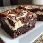 Two rich, fudgy squares of cream cheese brownies with a distinct white cream cheese swirl on top, served on a white plate.