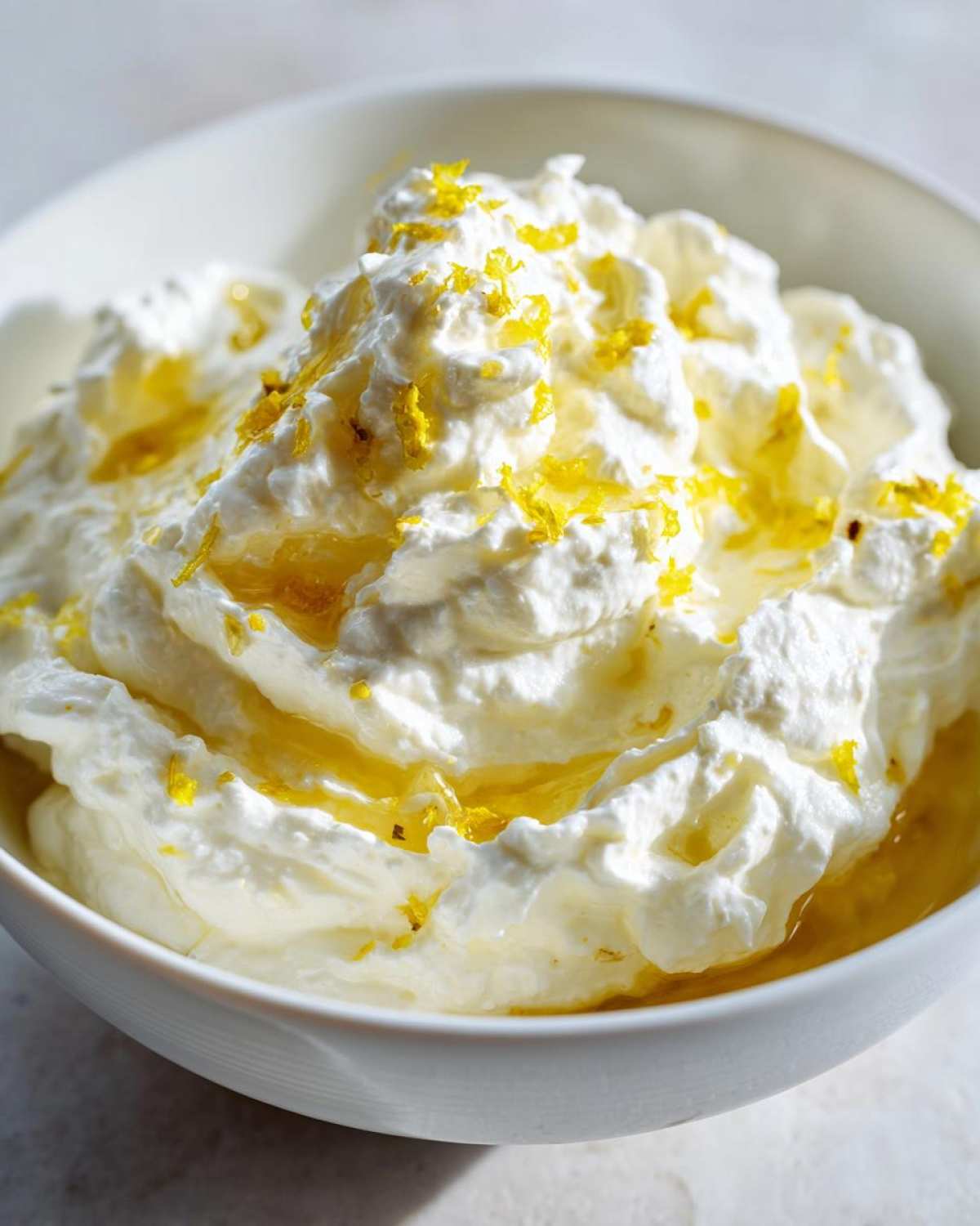 Close-up of creamy whipped feta dip swirled high in a white bowl, drizzled with olive oil and sprinkled with bright yellow lemon zest.