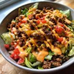 Close-up of a hearty cheeseburger bowl with ground beef, lettuce, tomatoes, pickles, melted cheese, and a drizzle of sauce.