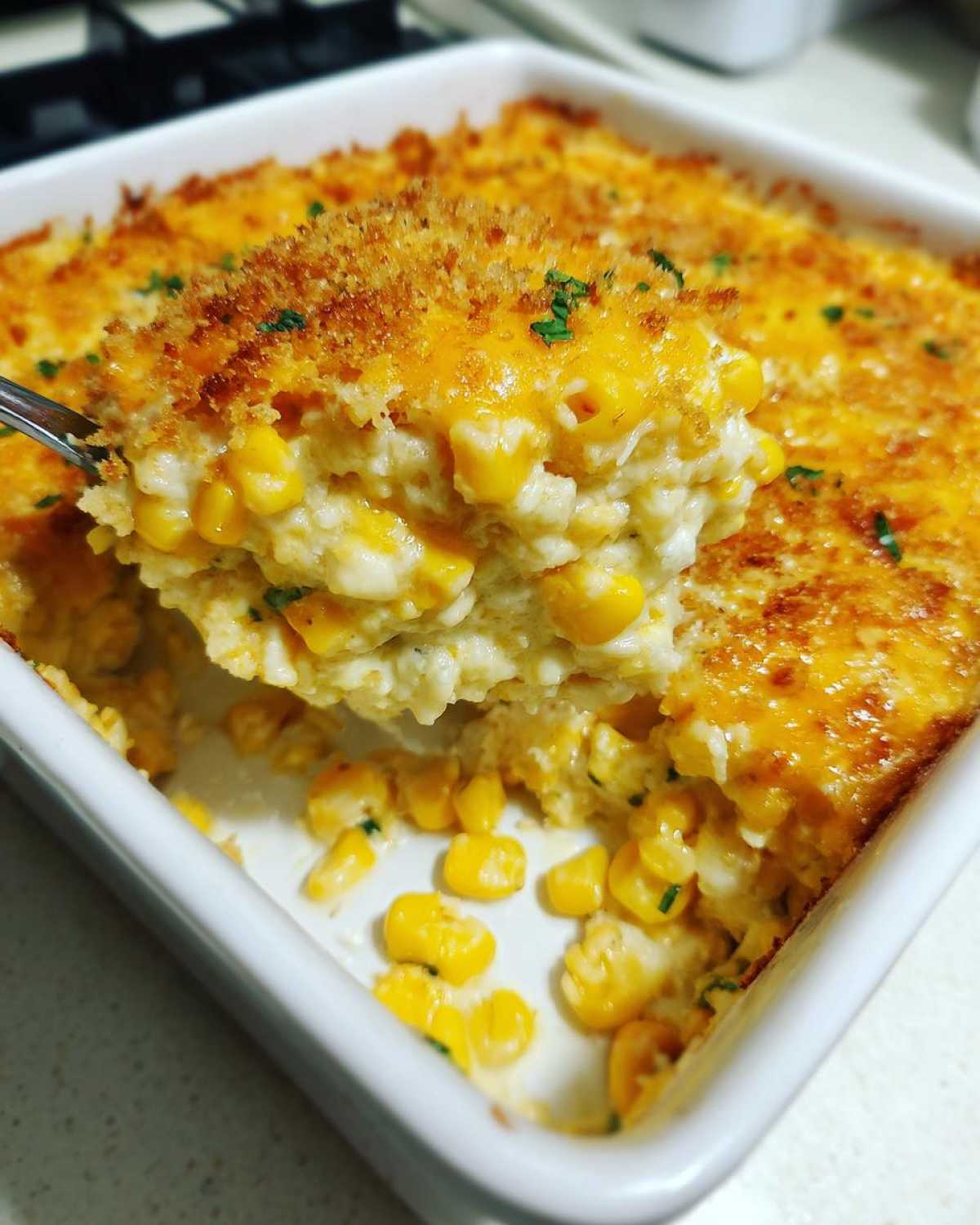 A spoonful of creamy, cheesy corn dip topped with golden breadcrumbs being lifted from a white baking dish.