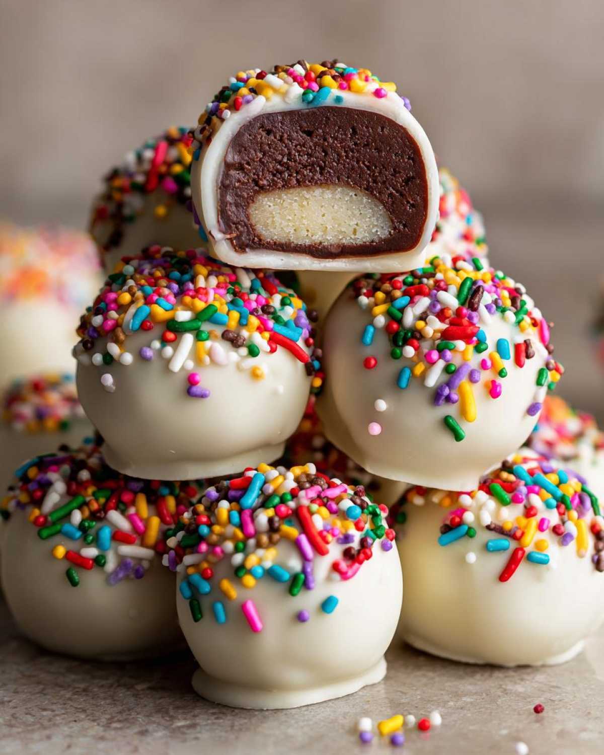 A stack of white chocolate coated sugar cookie truffles covered in rainbow sprinkles, one cut open.