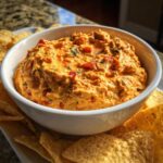 A close-up of a creamy, orange-colored boat dip mixed with red peppers and herbs, served in a white bowl surrounded by tortilla chips.