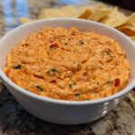 Close-up of a creamy, orange-hued boat dip mixed with red peppers and herbs, served in a white bowl with tortilla chips nearby.
