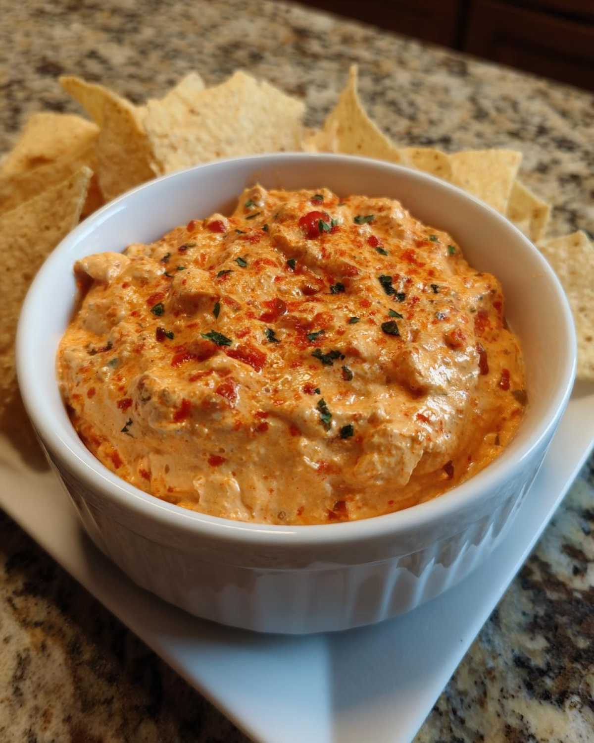 A creamy, orange-hued boat dip topped with red peppers and herbs, served with tortilla chips.