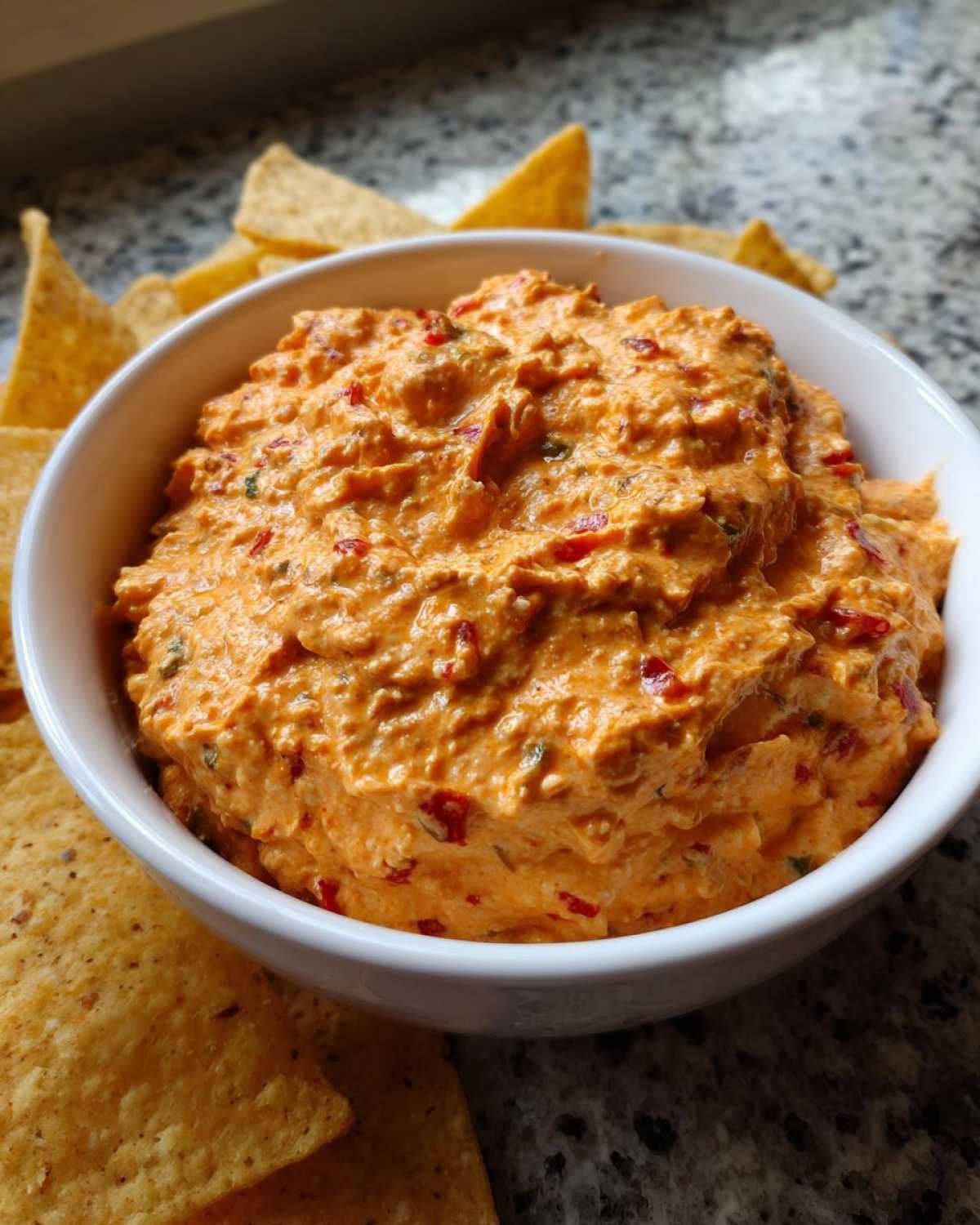 A white bowl filled with creamy, orange-colored boat dip mixed with red peppers, served with tortilla chips.
