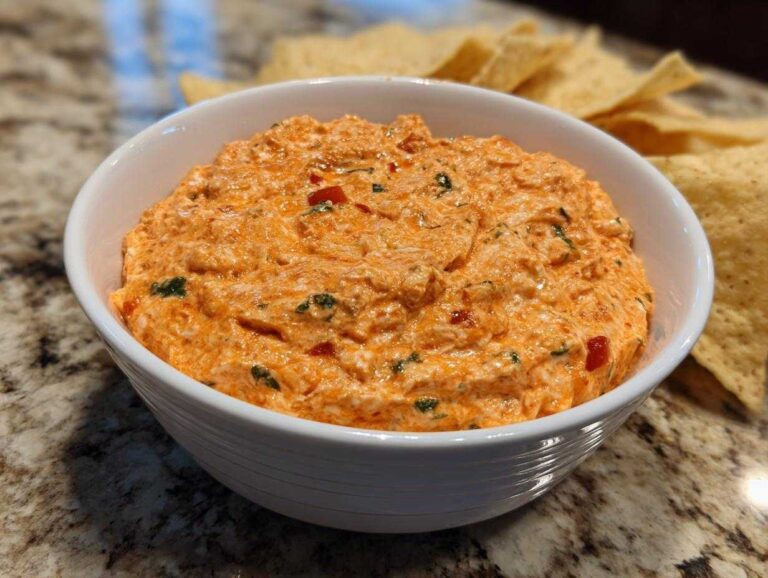 Close-up of a creamy, orange-hued boat dip mixed with red peppers and herbs, served in a white bowl with tortilla chips nearby.