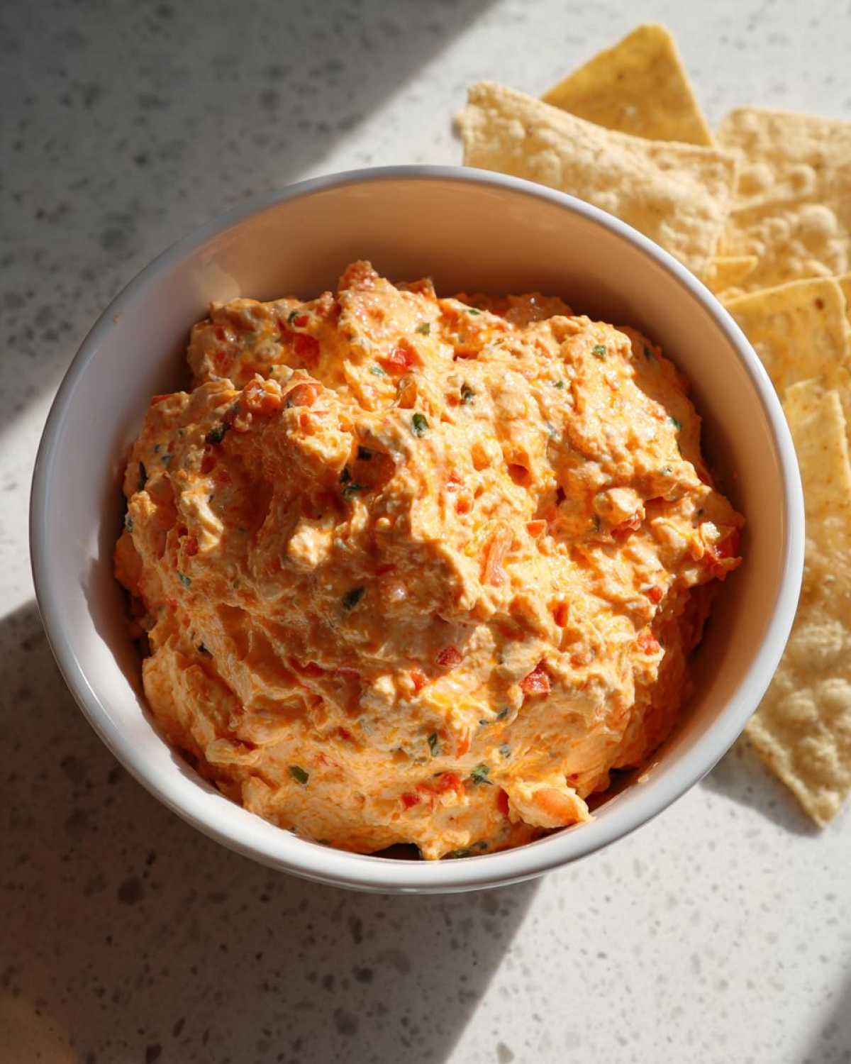 Overhead view of a creamy, orange-colored boat dip mixed with red peppers in a white bowl, served with tortilla chips.