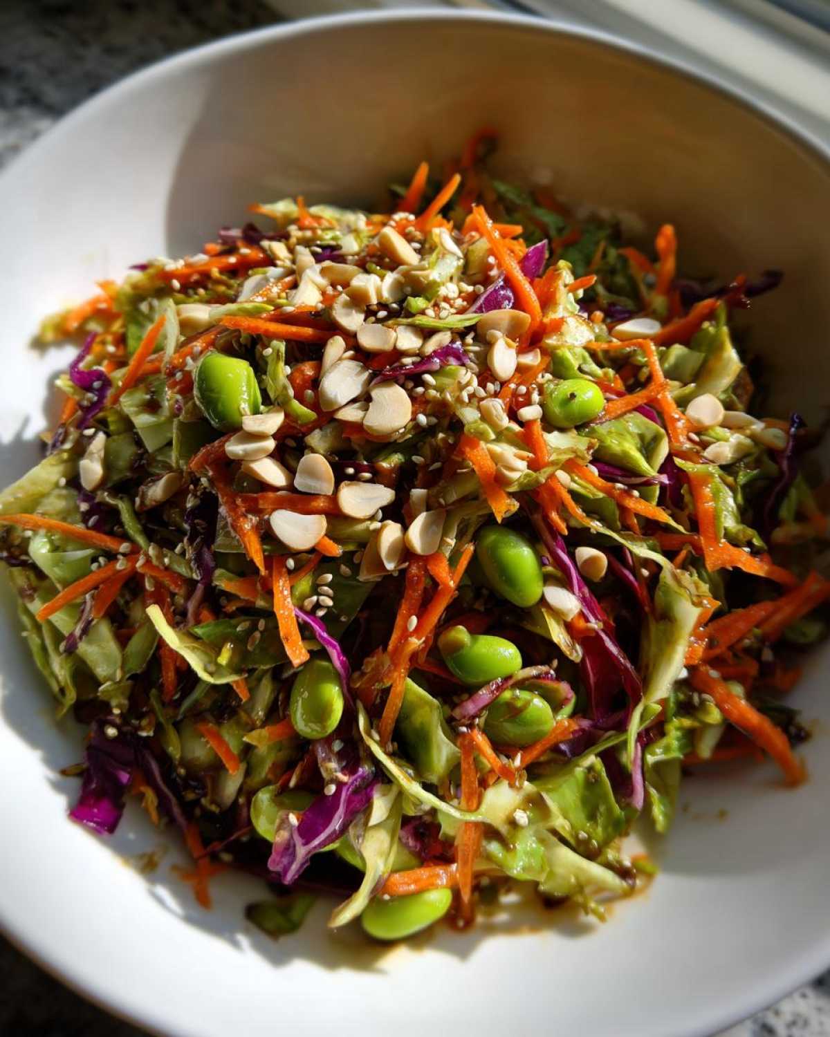 Close-up of a vibrant cabbage salad tossed in dressing, topped with shredded carrots, edamame, peanuts, and sesame seeds.