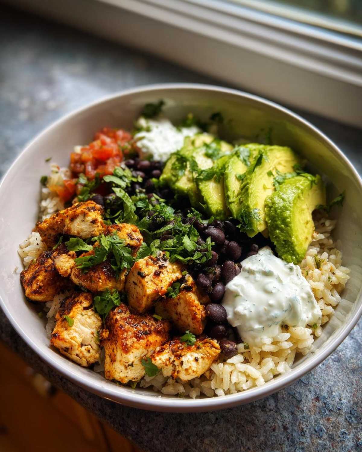 A vibrant chicken burrito bowl featuring seasoned chicken, rice, black beans, avocado slices, salsa, and sour cream.